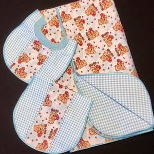 May include: A set of three baby bibs with a blue and white checkered pattern. The bibs are made of soft fabric and feature a cute teddy bear design.