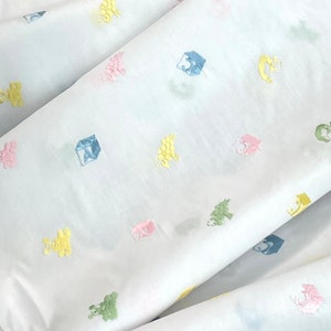 May include: White fabric with a repeating pattern of colourful embroidered toys. The toys include a rocking horse, a block, and a teddy bear.