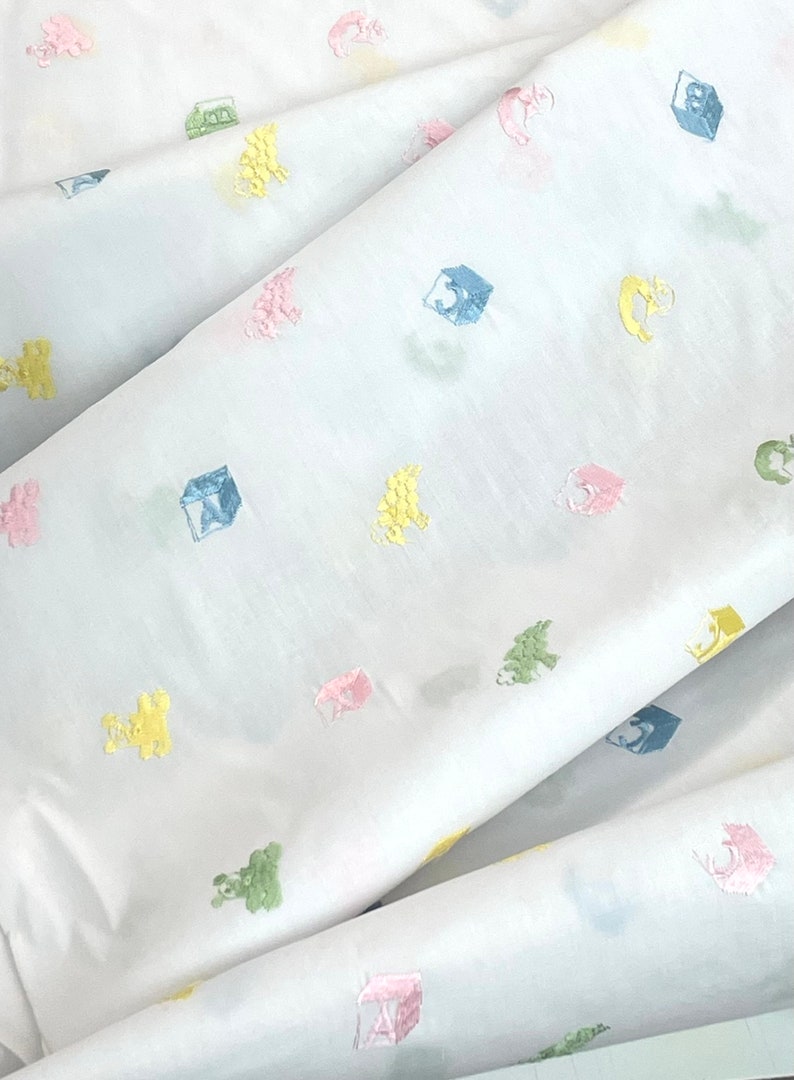 May include: White fabric with a repeating pattern of colourful embroidered toys. The toys include a rocking horse, a block, and a teddy bear.