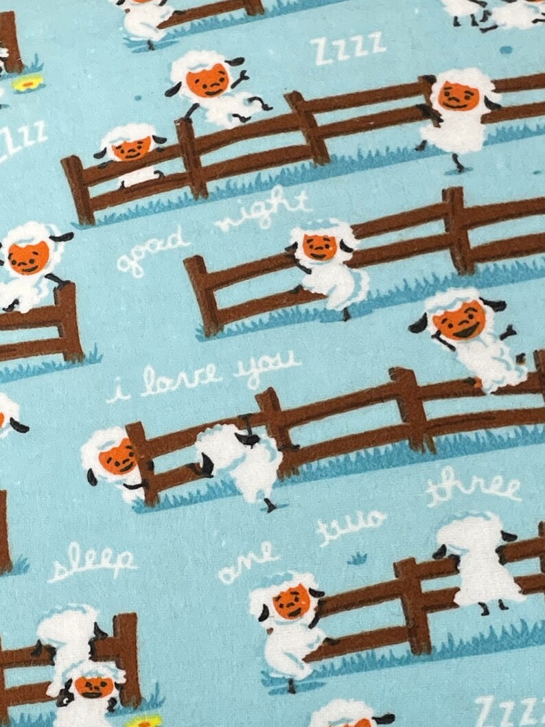 May include: A blue fabric with a repeating pattern of white sheep jumping over a brown fence. The text "good night", "i love you", "sleep", "one", "two", "three", and "zzz" is written on the fabric.