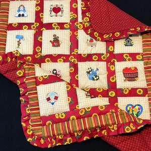 May include: A red and cream quilt with a sunflower pattern. The quilt is made up of squares with different embroidered designs, including a doll, a heart, a teapot, a watering can, a birdhouse, and a basket of apples. The quilt has a ruffled edge.