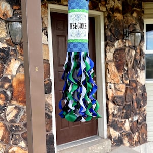 May include: A hanging welcome sign with blue, green, and white ribbons. The cylindrical sign has a blue and green patterned top and bottom, with the word "WELCOME" in the center. The sign is hanging in front of a brown door.