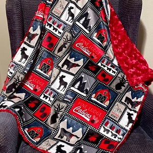 Puede incluir: Una manta de forro polar con estampado rojo y azul con respaldo de minky rojo. La manta presenta un diseño de patchwork con imágenes de alces, ciervos, árboles y cabañas. El texto "Pine Lodge Cabins" está impreso en la manta.