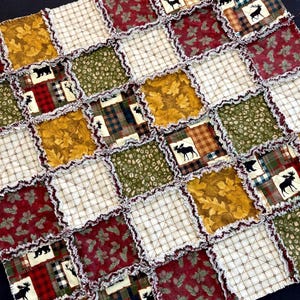May include: A patchwork quilt with a rustic theme. The quilt features squares of various fabrics in shades of red, gold, green, and cream. Some squares have leaf patterns, plaid, or bear and moose silhouettes. The edges are frayed for a cozy look.