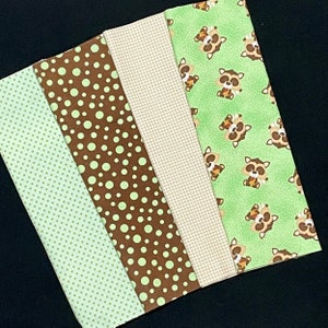 Peut inclure: Quatre carrés de tissu, l'un à pois marrons et verts, l'autre à carreaux marrons et blancs, l'un avec un fond vert et des ratons laveurs marrons, et l'autre à pois verts et blancs.