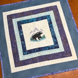 Peut inclure: Nappe carrée matelassée avec un motif d'ours et de montagne. La nappe présente un carré central avec une scène brodée d'ours et de montagne, entouré de carrés concentriques de motifs bleus et blancs variés. La bordure extérieure est bleue unie avec des pois blancs.