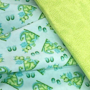 May include: A light blue fabric with a repeating pattern of green dragons with blue wings. The fabric is folded over a green fabric with white polka dots.