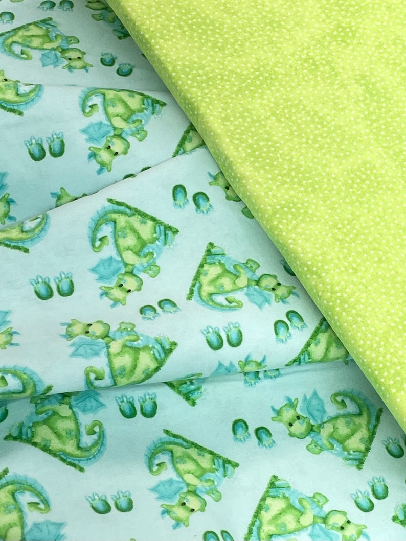 May include: A light blue fabric with a repeating pattern of green dragons with blue wings. The fabric is folded over a green fabric with white polka dots.