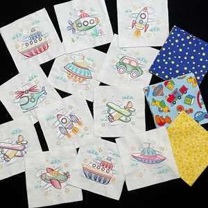 May include: A set of 12 fabric squares featuring embroidered images of various vehicles, including a spaceship, airplane, helicopter, car, submarine, and UFO. The squares are white with a light blue background and have a whimsical, cartoon-like style.