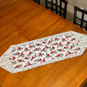 Könnte beinhalten: Ein weißer Tischläufer mit einem Muster aus roten Kardinalvögeln. Der Läufer hat einen grauen Rand mit einem Schneeflockenmuster. Der Läufer liegt auf einem Holztisch.