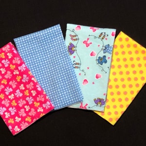May include: Four fabric squares in different patterns. One square is blue with a white checkered pattern. One square is pink with a white bow pattern. One square is yellow with a white polka dot pattern. One square is light blue with a white pattern of girls playing.