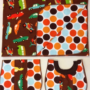 May include: A brown and white baby blanket with a car and truck pattern on one side and a polka dot pattern on the other side. The blanket is folded in half and there are two matching bibs with the same patterns.