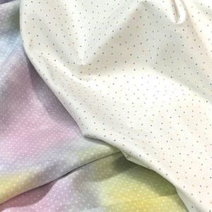 Può includere: Primo piano di tessuto bianco con un motivo a pois colorati. Il tessuto è piegato, rivelando un aspetto morbido e strutturato. Un secondo tessuto con una sfumatura arcobaleno pastello è visibile.