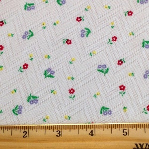 K&ouml;nnte beinhalten: Wei&szlig;er Stoff mit einem kleinen, sich wiederholenden Muster aus roten, gelben, gr&uuml;nen und violetten Blumen. Ein Holzliniall mit Zollmarkierungen liegt auf dem Stoff.