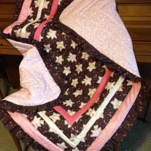 May include: A pink and brown baby quilt with a star pattern and white hearts. The quilt has a ruffled edge and is made of soft fabric.