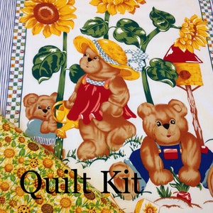 May include: Quilt kit featuring a white background with a whimsical design of three teddy bears surrounded by sunflowers and green leaves. The text "Quilt Kit" is printed in black on the fabric.