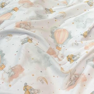 May include: White fabric with a whimsical pattern featuring bunnies, hot air balloons, moons, and stars in soft pastel colors. The design includes peach, gray, and yellow accents, creating a dreamy, celestial theme.