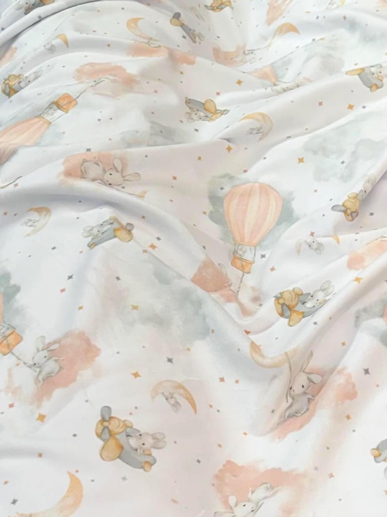 May include: White fabric with a whimsical pattern featuring bunnies, hot air balloons, moons, and stars in soft pastel colors. The design includes peach, gray, and yellow accents, creating a dreamy, celestial theme.