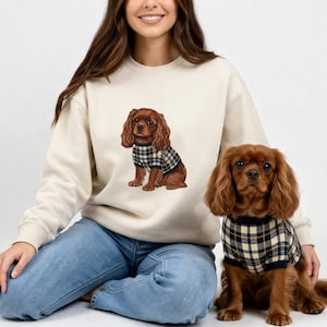 Può includere: Una felpa color crema con un'illustrazione di un Cavalier King Charles Spaniel con un maglione a quadri. Un vero cane Cavalier King Charles Spaniel con un maglione a quadri abbinato è seduto accanto alla persona che indossa la felpa. Il cane ha lunghi peli marroni.
