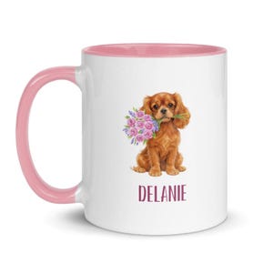Pode incluir: Caneca de cerâmica branca com interior e alça rosa. A caneca apresenta uma ilustração em aquarela de um Cavalier King Charles Spaniel segurando um buquê de flores rosa e roxas. O nome "DELANIE" é impresso em bordô.