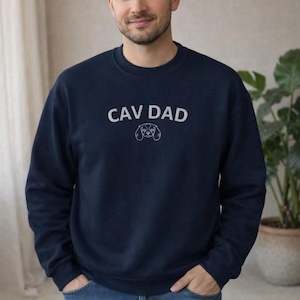 Op de afbeelding: Marineblauwe sweatshirt met de witte tekst "CAV DAD" en een witte lijntekening van een Cavalier King Charles Spaniel-gezicht. Ronde hals.