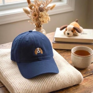 Puede incluir: Gorra de béisbol azul marino con un pequeño bordado de la cara de un perro en la parte delantera. La gorra está sobre una manta de punto color crema. Un juguete de perro de peluche y una taza de té también están en la imagen.