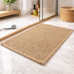 May include: A rectangular, beige woven bath mat with a fringed edge. The mat has a grid-like pattern and is placed on a marble floor. The bathroom setting includes a shower head, wooden accents, and a wicker basket.