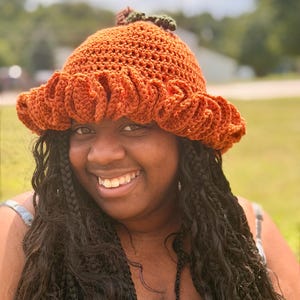 May include: A burnt orange crochet bucket hat featuring a wide, ruffled brim and a small, dark green stem detail at the top center. The hat is constructed with a textured, ribbed stitch pattern throughout the crown and the flared, wavy brim.