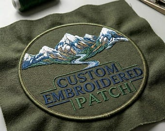 May include: A circular embroidered patch on olive green fabric featuring a mountain landscape with snow-capped peaks and a winding river. The text "CUSTOM EMBROIDERED PATCH" is stitched in dark blue and green thread below the mountain design.