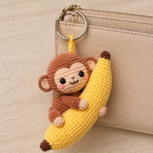 Op de afbeelding: Gehaakte aap sleutelhanger die een gele banaan omhelst. De aap is bruin met een lichtbruin gezicht, roze wangen en zwarte ogen. De banaan heeft een bruine punt. De sleutelhanger is bevestigd aan een zilveren ring.