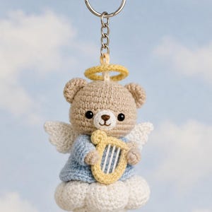 May include: A crocheted teddy bear keychain with angel wings, a halo, and a harp. The bear is light brown, wearing a blue shirt, and sitting on a white cloud. The keychain has a silver ring.
