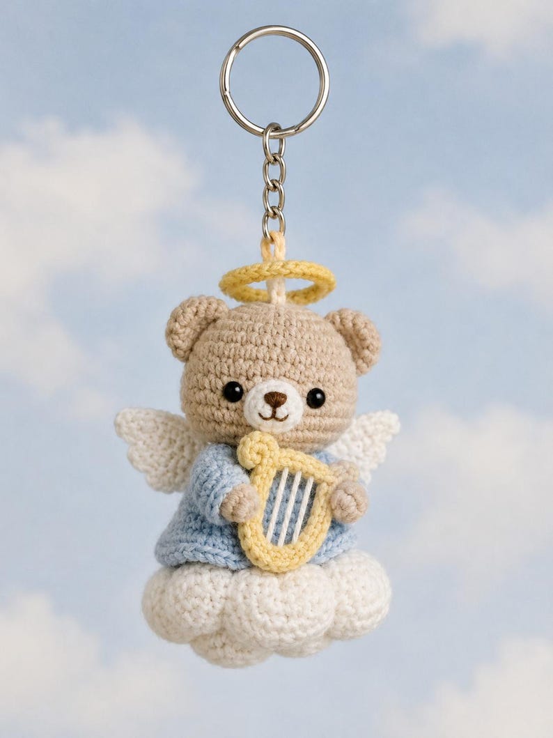 May include: A crocheted teddy bear keychain with angel wings, a halo, and a harp. The bear is light brown, wearing a blue shirt, and sitting on a white cloud. The keychain has a silver ring.