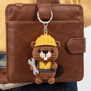 May include: A crocheted beaver keychain with a yellow hard hat, overalls, and a wrench. The brown beaver has a white muzzle and a dark brown tail. The keychain is attached to a silver ring and hangs in front of a brown leather wallet.