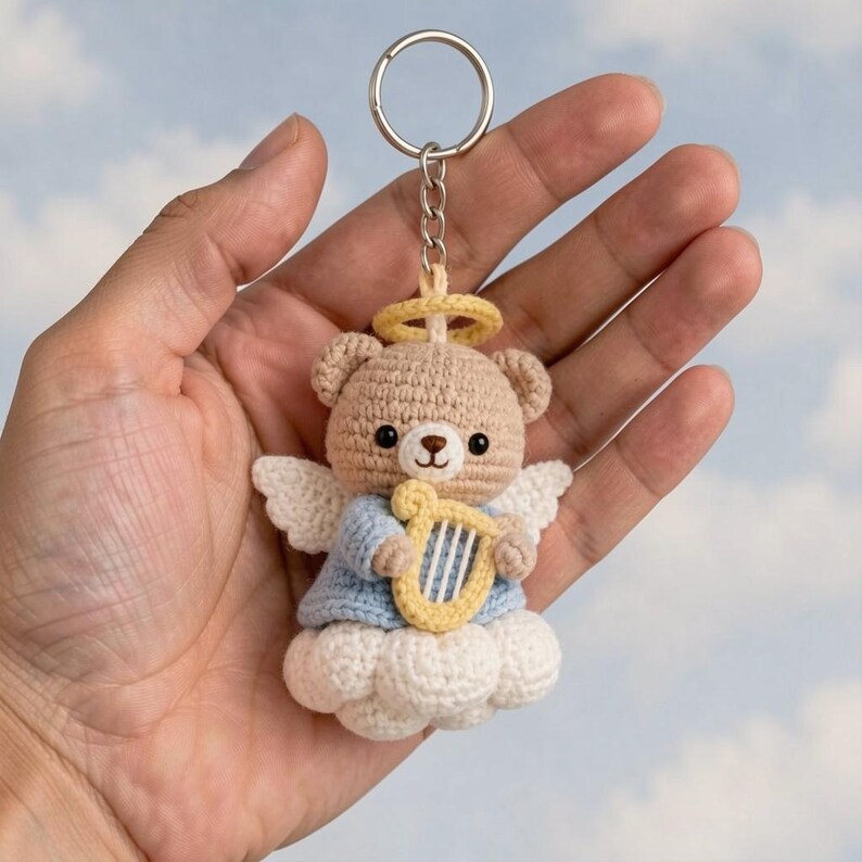 May include: A crocheted angel bear keychain. The bear is tan with a yellow halo, white wings, and a blue shirt. It holds a yellow harp and sits on a white cloud. The keychain is attached to a silver ring.