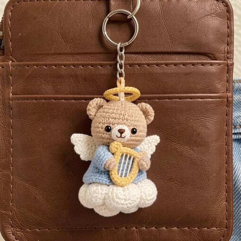 May include: Crocheted angel bear keychain. The bear is light brown with white wings, a blue shirt, and a yellow harp. It has a yellow halo and sits on a white cloud. The keychain is attached to a silver ring.