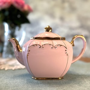 Puede incluir: Una tetera de cerámica rosa con adornos y detalles dorados. La tetera tiene un pico y un asa curvos, y una tapa a juego con un adorno dorado. Líneas y espirales doradas decorativas adornan el cuerpo. Un artículo clásico para la hora del té.