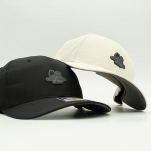 May include: Two baseball caps, one black and one off-white, are displayed. Each cap features a curved brim and a dark gray patch with the words "CAT MOM" and a cat silhouette. The caps are angled, showcasing their design.