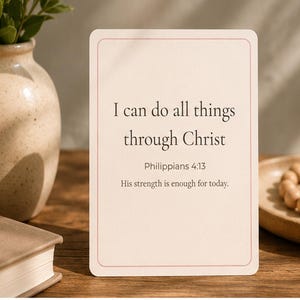 May include: A cream-colored card with the text "I can do all things through Christ" and the scripture reference Philippians 4:13. The card is on a wooden surface, next to a vase with greenery and a book.