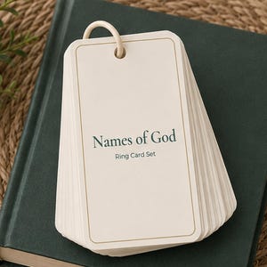 May include: A stack of off-white rectangular cards held together by a light-colored plastic ring. The top card features the dark green text "Names of God" and "Ring Card Set" with a thin gold border surrounding the edge of the card.