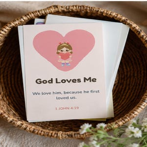 May include: A rectangular card featuring a pink heart graphic with a cartoon character holding a smaller heart. The text reads God Loves Me, We love him, because he first loved us, and 1 JOHN 4:19. The card is placed inside a woven brown basket.