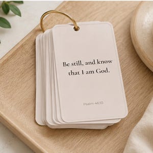 May include: A stack of rectangular off-white cards held together by a gold metal ring. The top card features the black printed text, Be still, and know that I am God, followed by the smaller text, Psalm 46:10, centered on the card surface.