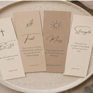 May include: Four rectangular paper bookmarks in shades of beige and tan. Each features black text and minimalist line art icons, including a cross, a leaf sprig, and a sun. The bookmarks display religious phrases and scripture citations.
