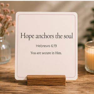 以下が含まれることがあります： 「Hope anchors the soul」のテキストと聖書の詩が書かれた、角が丸い正方形のカード。カードは小さな木のブロックで支えられています。ガラスの瓶に入った火の灯ったろうそくと、ドライフラワーの花瓶が背景にあります。