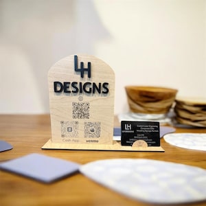 May include: A wooden sign with the text "LH DESIGNS" in black, featuring QR codes for Cash App and Venmo. A business card with contact information sits in front of the sign. The sign is on a wooden table.