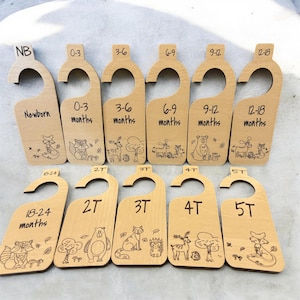 May include: Wooden baby clothes hangers with engraved designs and age labels: Newborn, 0-3 months, 3-6 months, 6-9 months, 9-12 months, 12-18 months, 18-24 months, 2T, 3T, 4T, and 5T. Each hanger features a different woodland animal illustration.