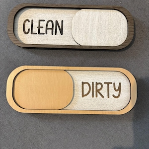 Może przedstawiać: Dwa owalne drewniane magnesy na zmywarkę z przesuwanymi panelami. Górny magnes ma ramkę z ciemnego drewna i wyświetla słowo CLEAN czarną czcionką. Dolny magnes ma ramkę z jasnego drewna i wyświetla słowo DIRTY czarną czcionką.