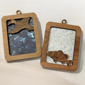 May include: Two rectangular wooden frames with hanging loops. One frame features a laser-cut wooden duck silhouette over a dark blue bokeh background. The other frame displays a laser-cut wooden fish silhouette over a white floral pattern background.