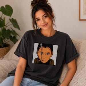 Pu&ograve; includere: T-shirt grigio scuro con una grafica quadrata di un personaggio anime con una sigaretta. Il personaggio ha i capelli scuri, una cicatrice e un'espressione seria. La maglietta ha maniche corte e una vestibilit&agrave; rilassata.