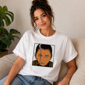 Pu&ograve; includere: T-shirt bianco con un'illustrazione in stile anime incorniciata di una persona con una sigaretta. L'illustrazione utilizza tonalit&agrave; marroni, nere e bianche. La t-shirt ha maniche corte e girocollo.