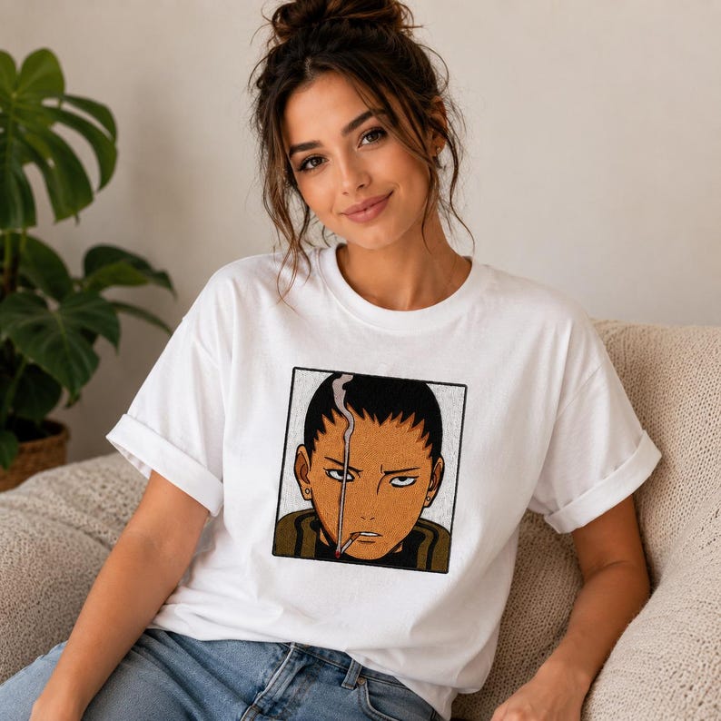 Pu&ograve; includere: T-shirt bianco con un'illustrazione in stile anime incorniciata di una persona con una sigaretta. L'illustrazione utilizza tonalit&agrave; marroni, nere e bianche. La t-shirt ha maniche corte e girocollo.